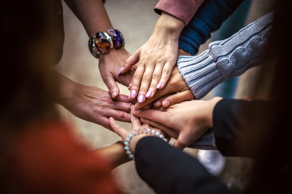A group of diverse hands stacked together in a show of unity and community, with various bracelets and clothing styles visible.