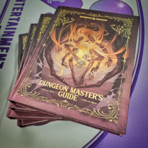 A stack of the 2024 Dungeon Master’s Guide books, each featuring a cover illustration of an eerie spider-like figure with flowing hair, hovering over a victim wrapped in webbing. The cover has intricate gold filigree detailing, with the title "Dungeon Master's Guide" in bold, gold lettering at the bottom. The books are stacked on a surface with purple and grey tones in the background.