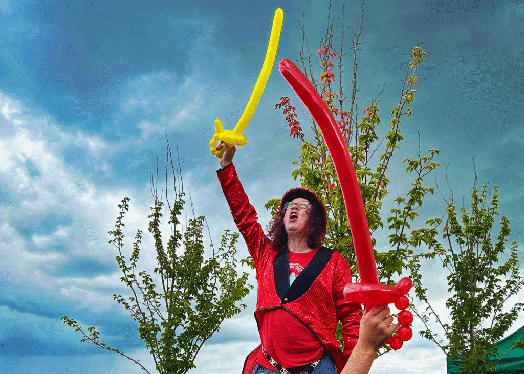 Performer on stilts in a red sequinned jacket holding balloon swords at a Dice and Balls CIC community event in Nottingham.