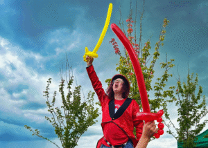 Performer on stilts in a red sequinned jacket holding balloon swords at a Dice and Balls CIC community event in Nottingham.